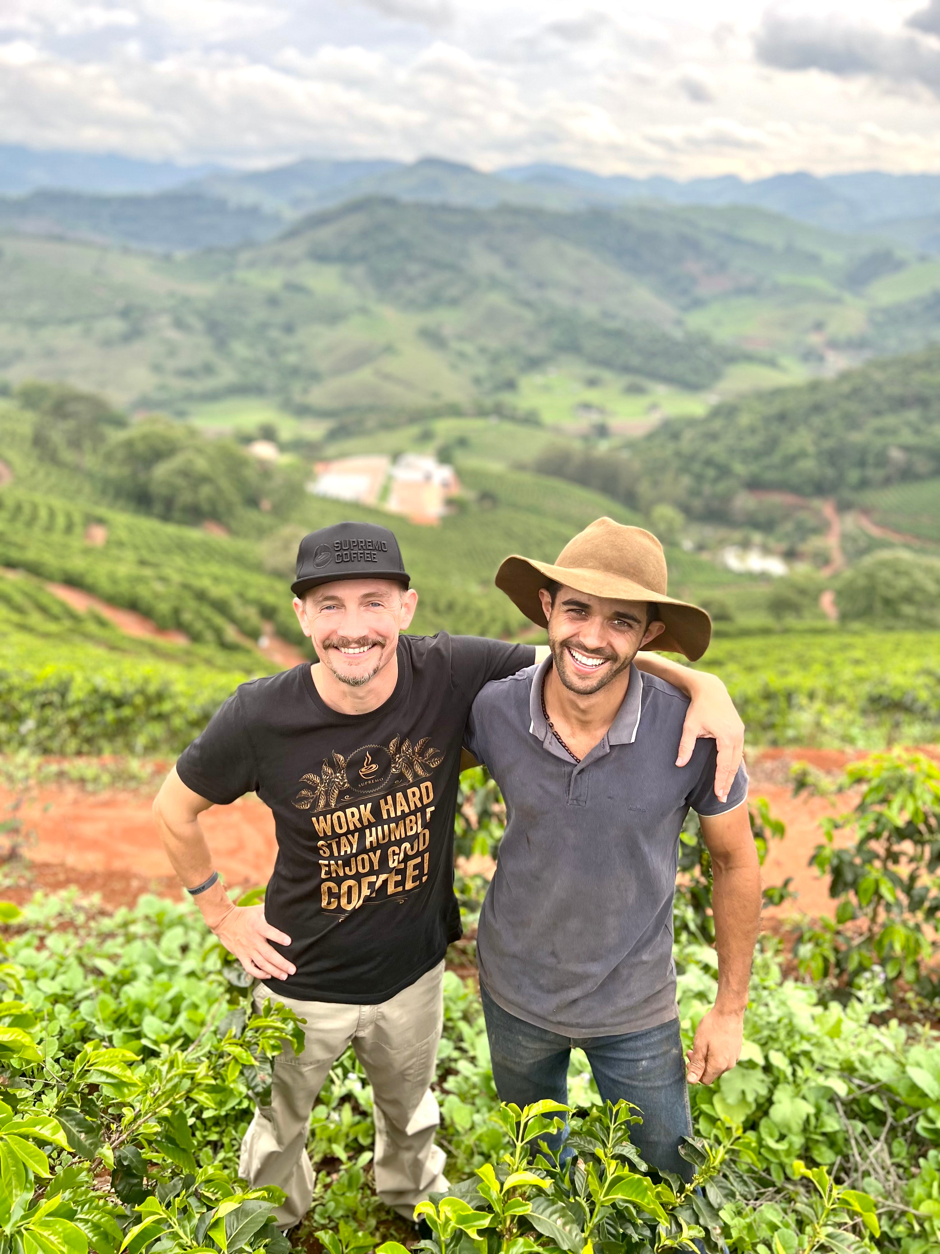 Antonio Marcio da Silva und Raphael Braune (SUPREMO) Arm in Arm auf einer Kaffeeplantage. Froschperspektive und Blick ins Tal im Hintergrund. Sítio Baixadão, Brasilien | SUPREMO Coffee